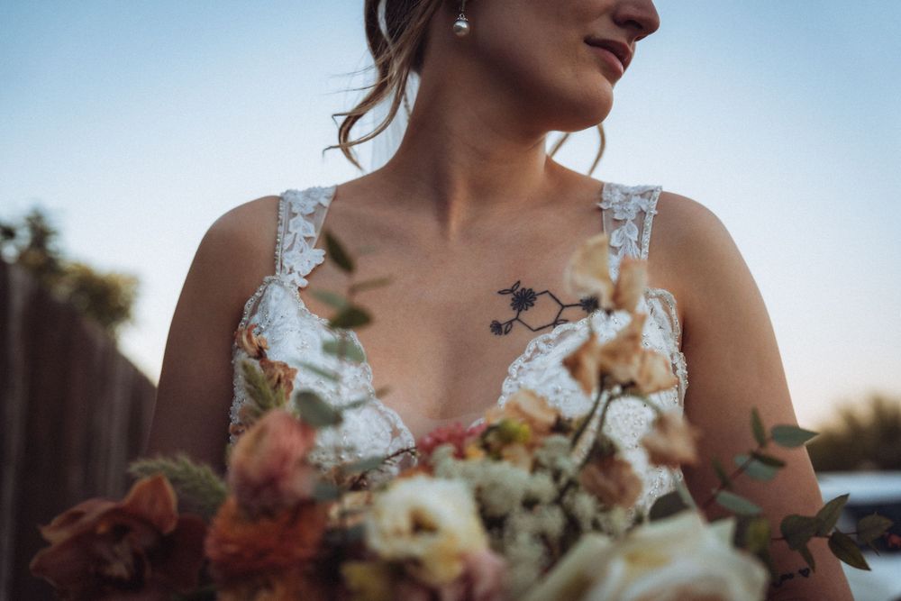 Bride in lace wedding dress with floral chest tattoo holding a bouquet of pink and white flowers at sunset.