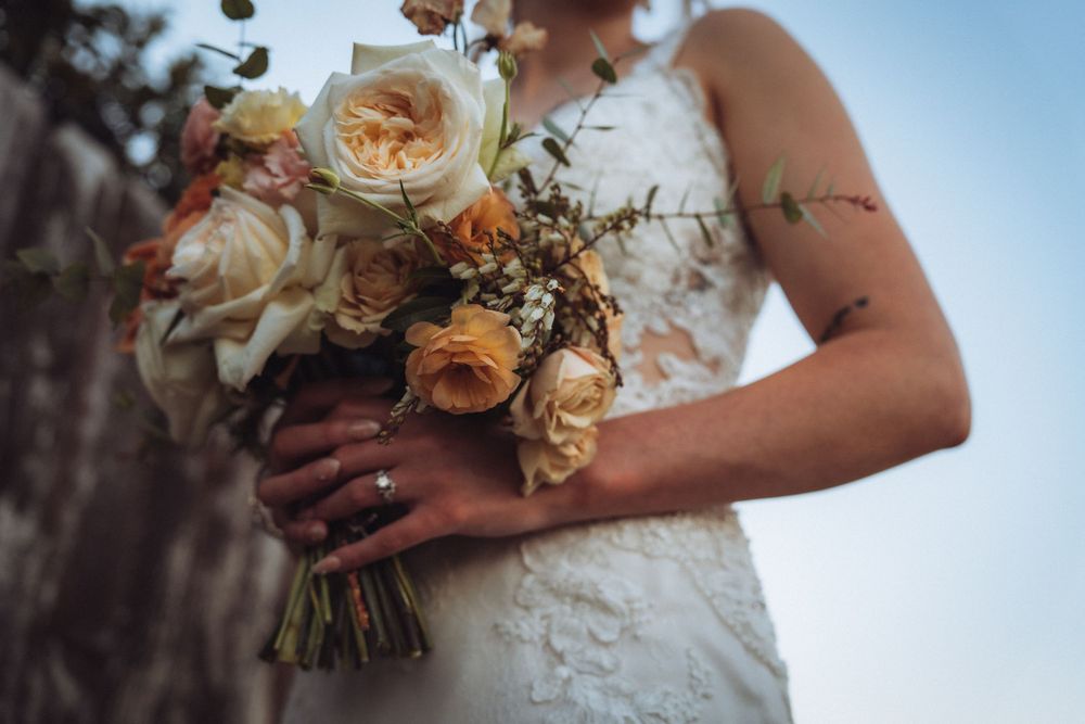 Bride in lace wedding dress holding elegant bouquet of peach and cream roses with greenery.
