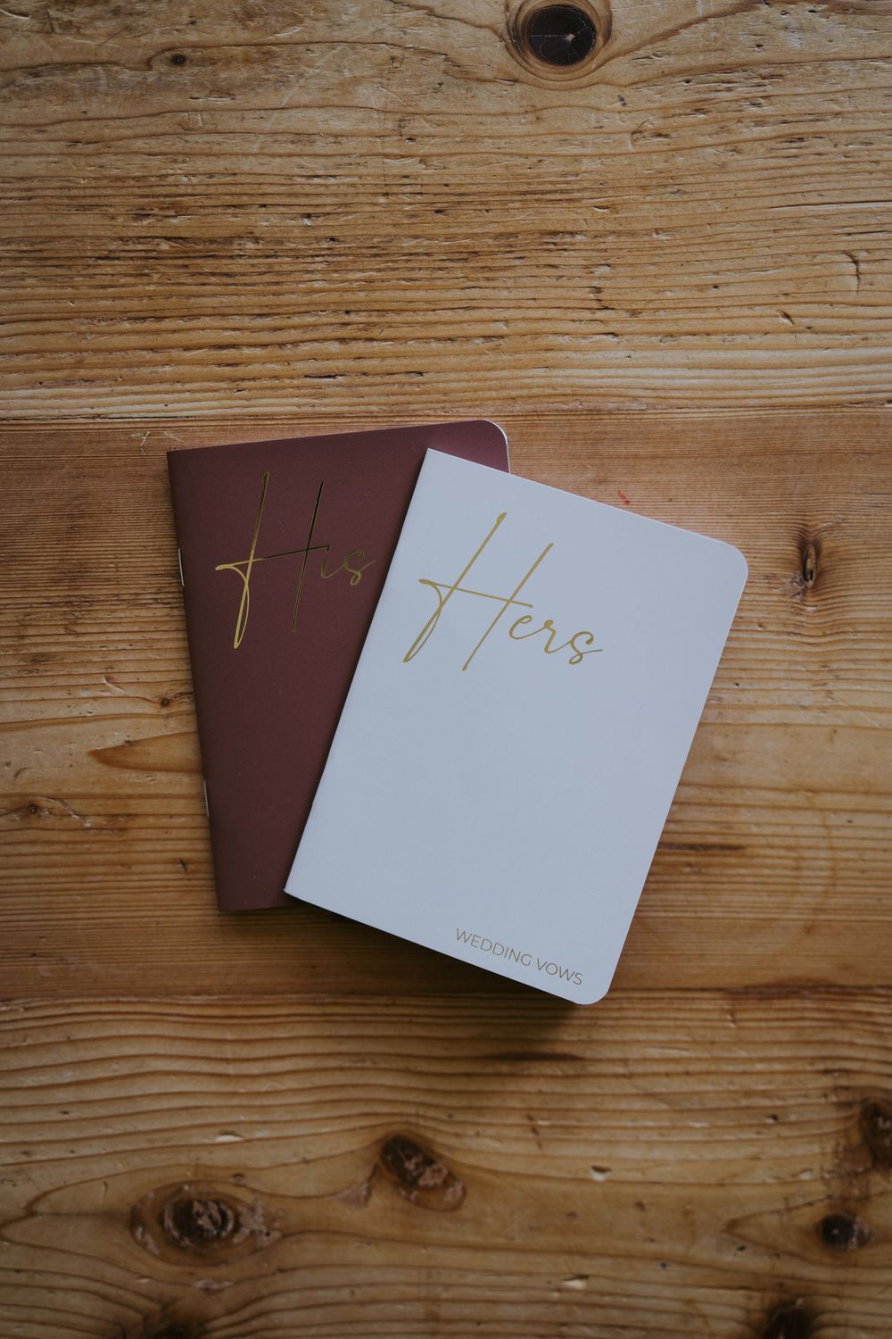 His and Hers wedding vow books in burgundy and white with gold lettering on a rustic wooden surface.