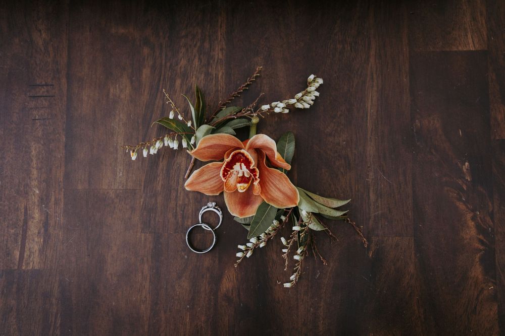 Orange orchid boutonniere with greenery and wedding rings on dark wood surface.