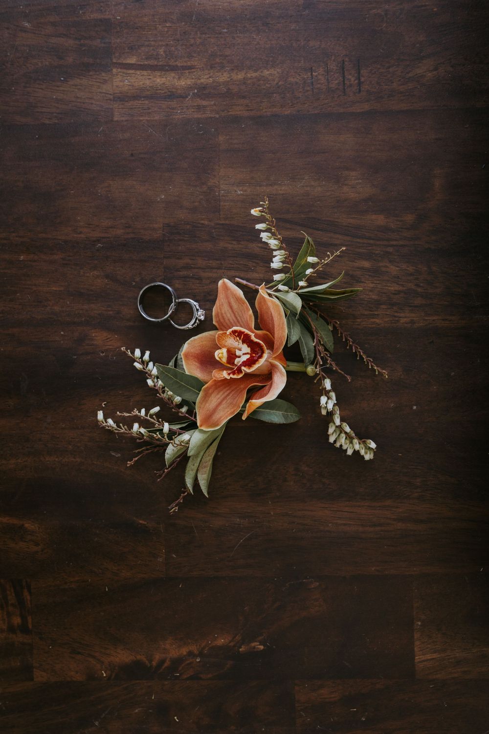 Wedding rings resting on an orange cymbidium orchid boutonniere with greenery on a dark wood surface.
