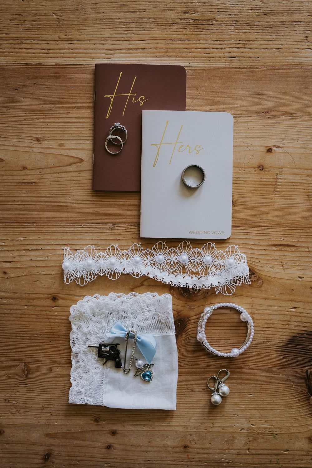 Wedding flat lay with His and Hers vow books, gold rings, lace garter, pearl earrings, and bridal accessories on wood.