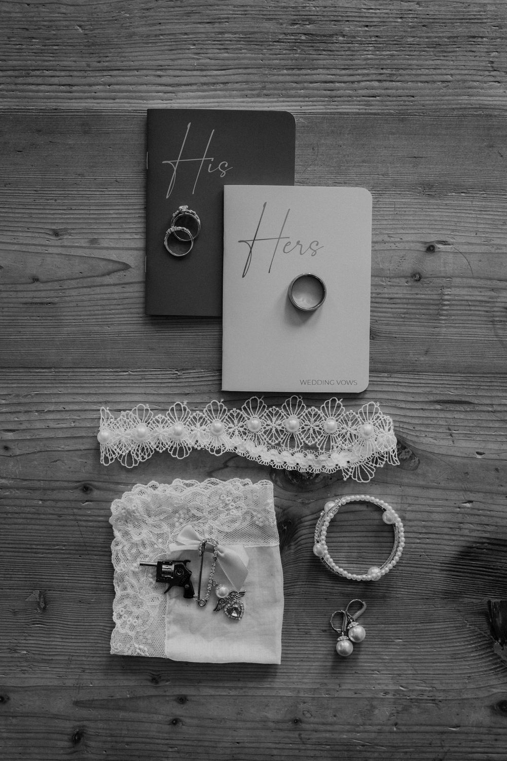Black and white flat lay of wedding vow books, rings, lace garter, bracelet, and pearl earrings on wood.