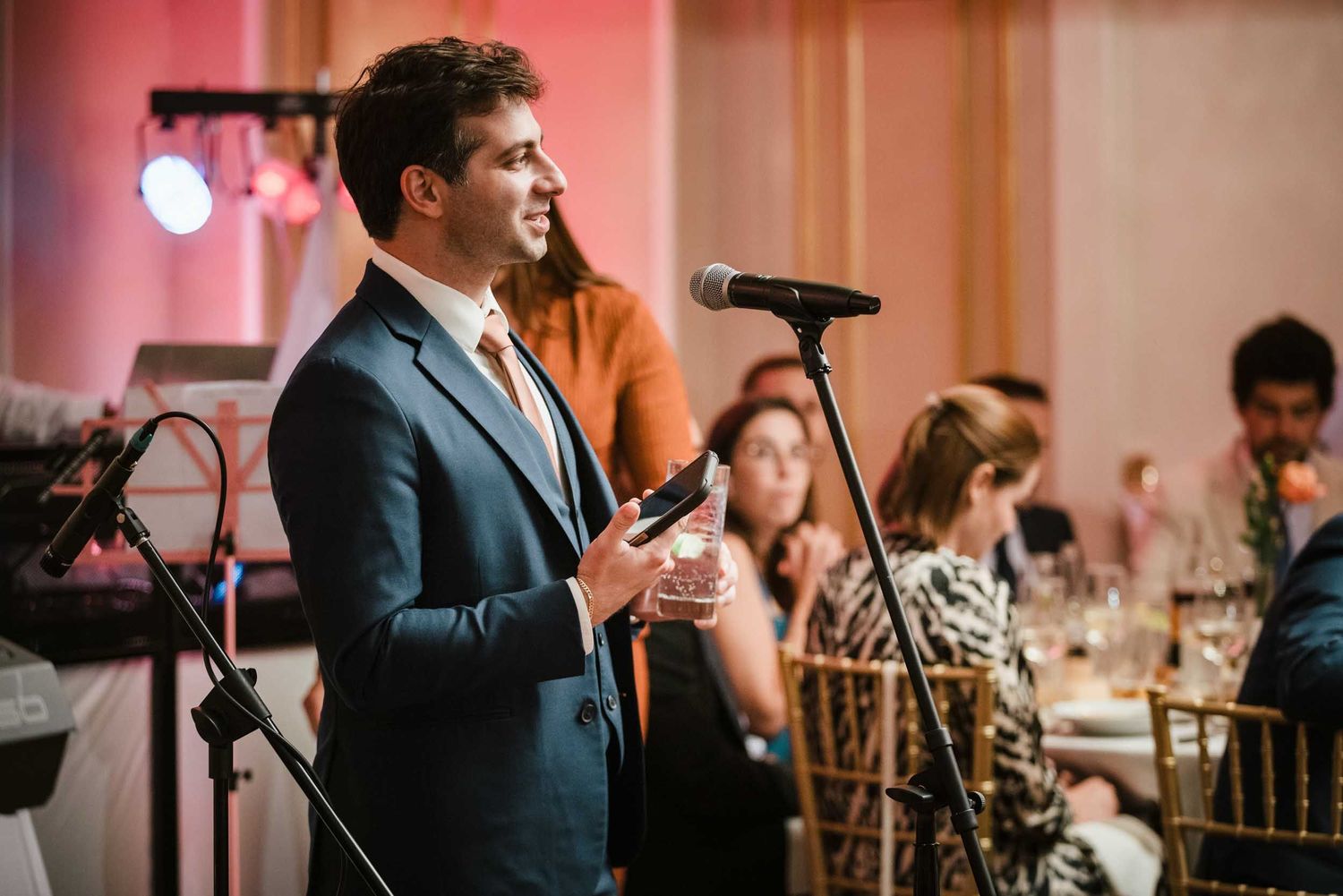 The best man giving a speech while holding a drink at the microphone during a Boston Lenox Hotel wedding reception