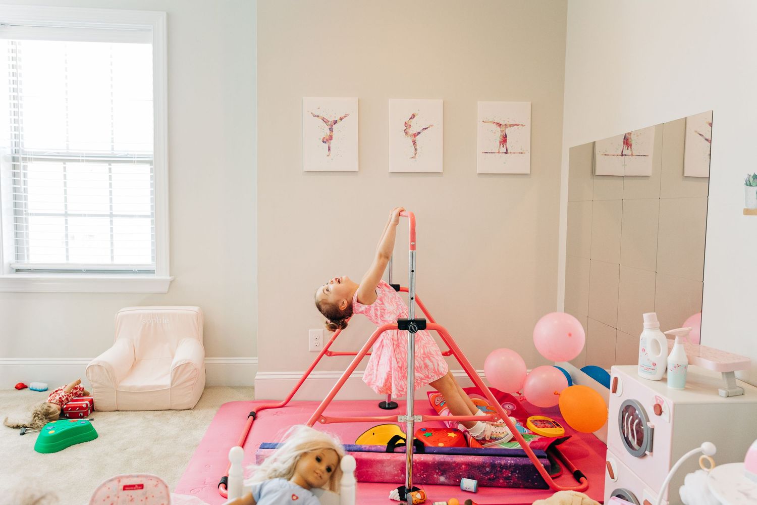 A pink gymnastics mat and play area setup in a bright room with ballet artwork on the walls and scattered toys with a little girl playing