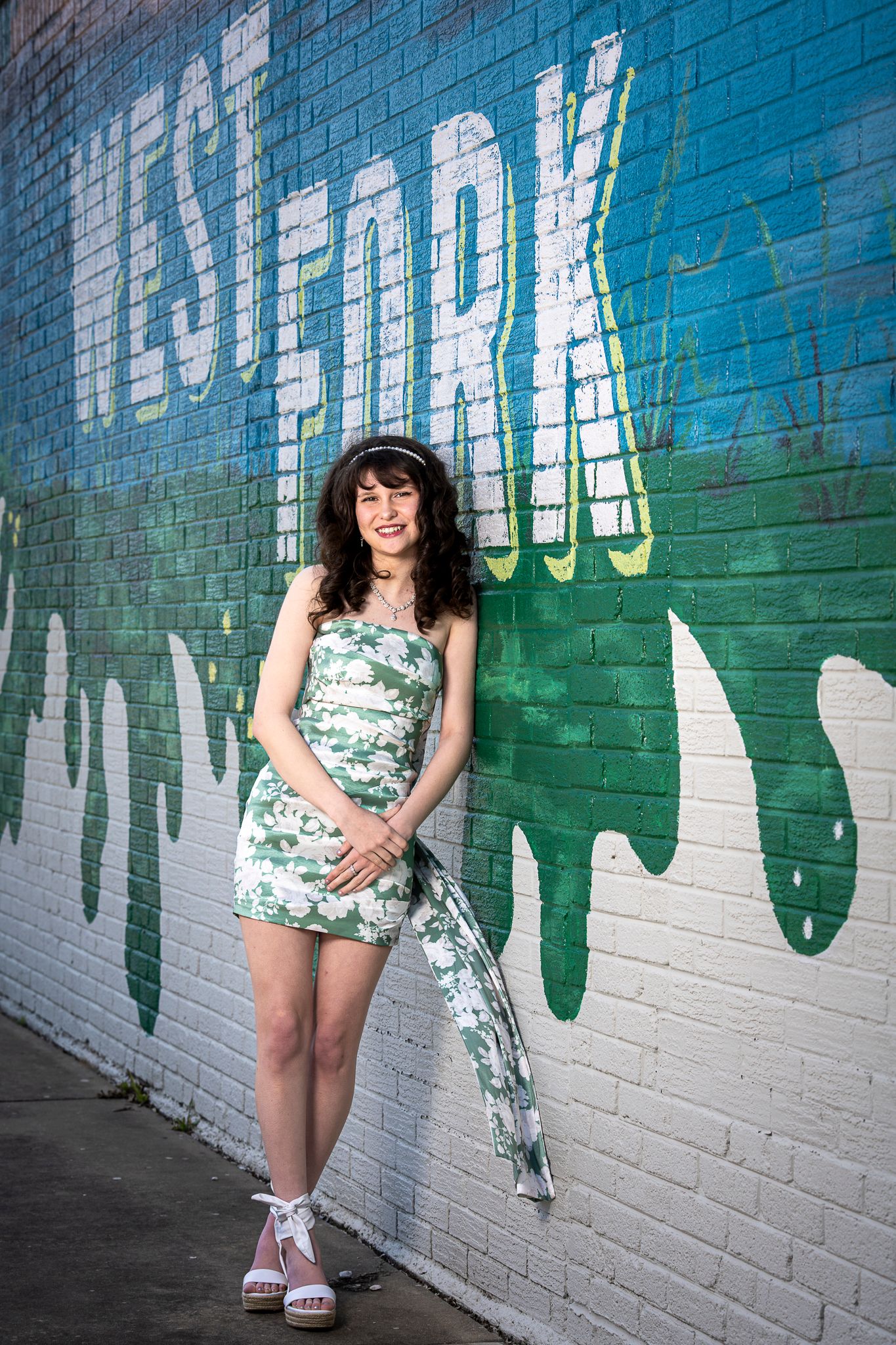 A person in a floral dress and black jacket poses against a blue and green graffiti wall with a metal railing.