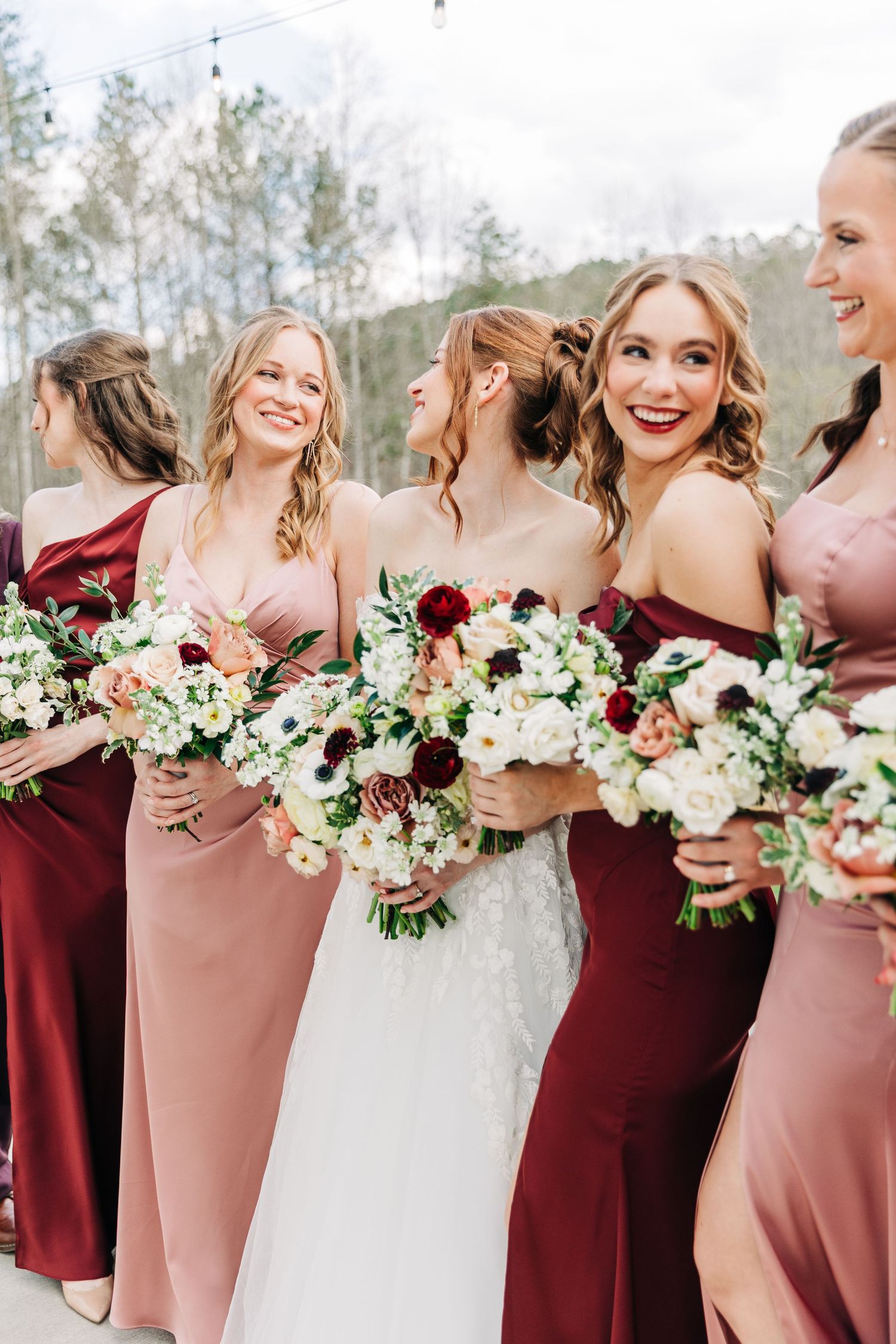 Wedding party in alternating burgundy and blush pink dresses hold white and red floral bouquets while posing together.