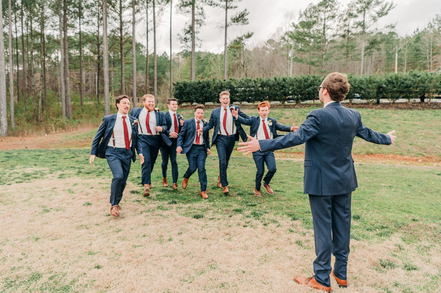 A group of people in formal attire gather outdoors in a grassy field near a pine forest.