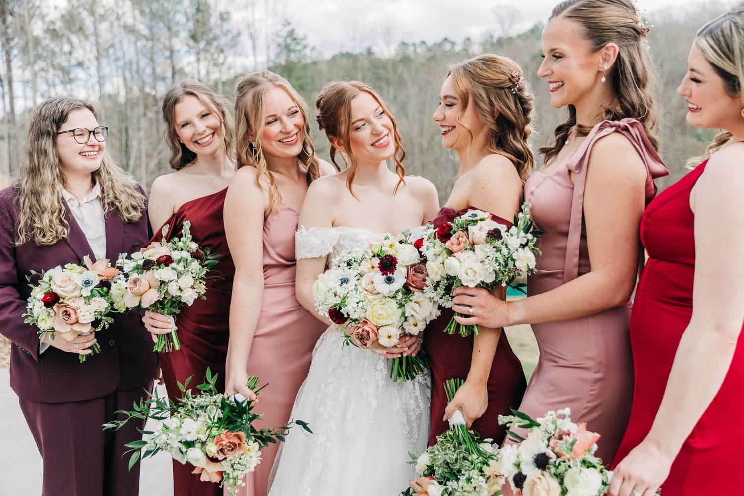 Wedding party wearing pink and burgundy dresses pose outdoors with floral bouquets in winter setting.