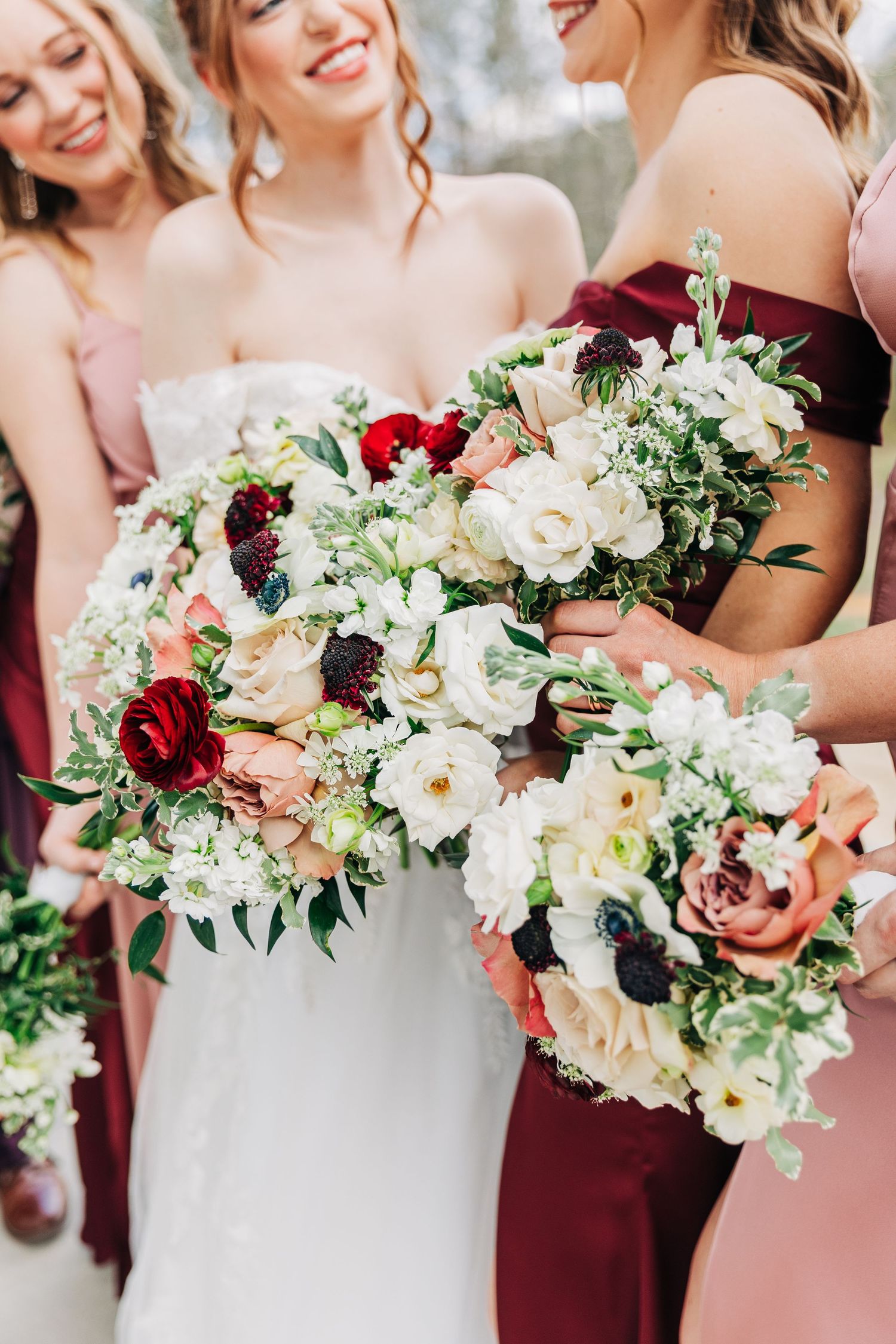 Bridesmaids holding elegant bouquets of white and burgundy flowers with greenery at a wedding celebration.