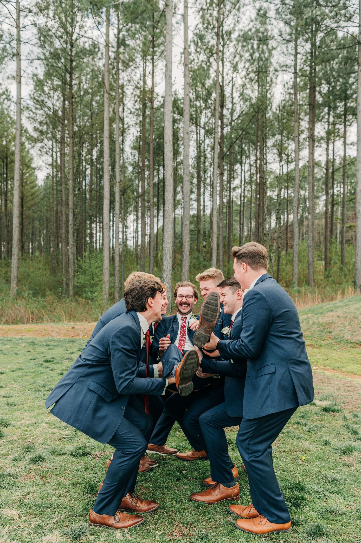 Groomsmen in navy suits and brown shoes playfully pose in sequence on grassy woodland ground.