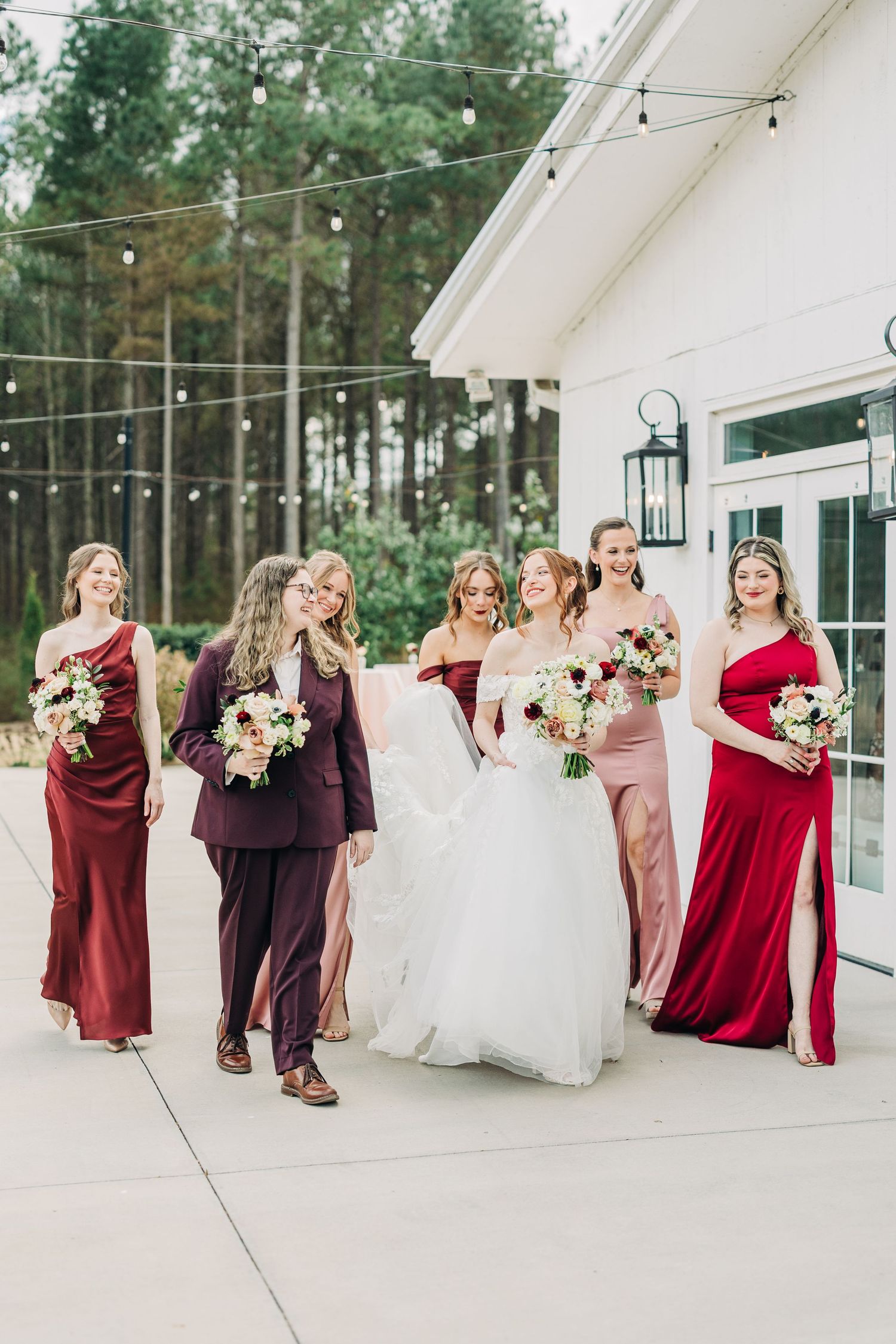 Bridal party in burgundy and blush dresses laugh together while walking outside a white building.