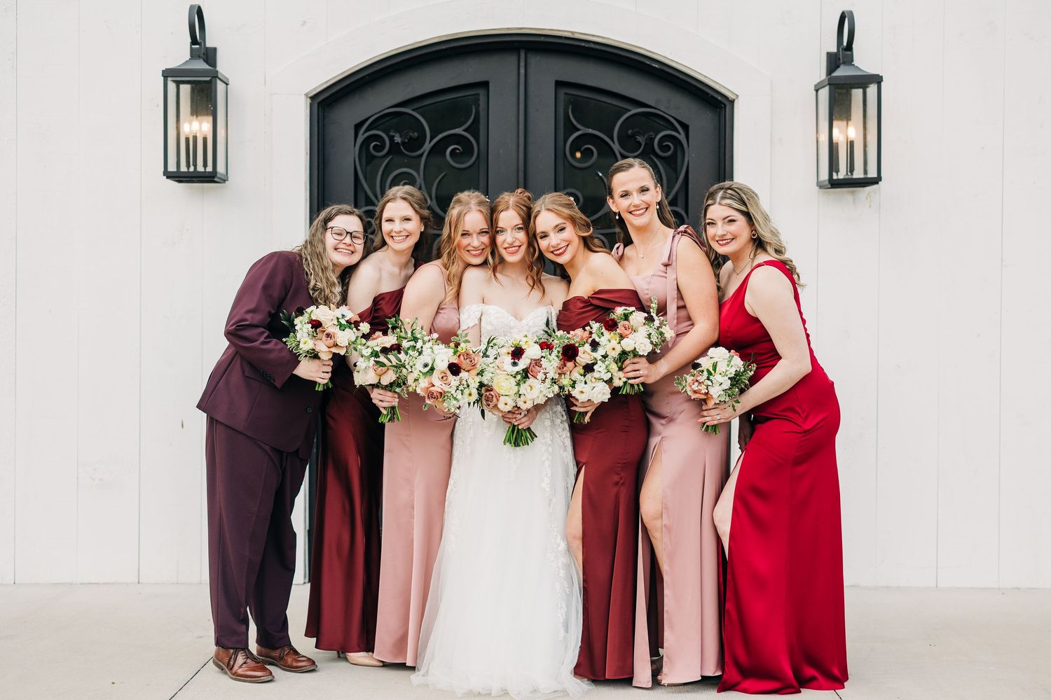 Bridal party in blush pink and burgundy dresses stand together holding white and pink bouquets.