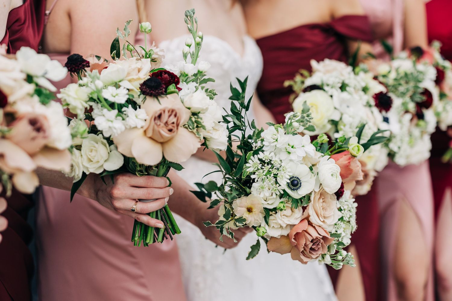 Row of bridesmaids holding elegant white and blush pink floral bouquets with roses and anemones.