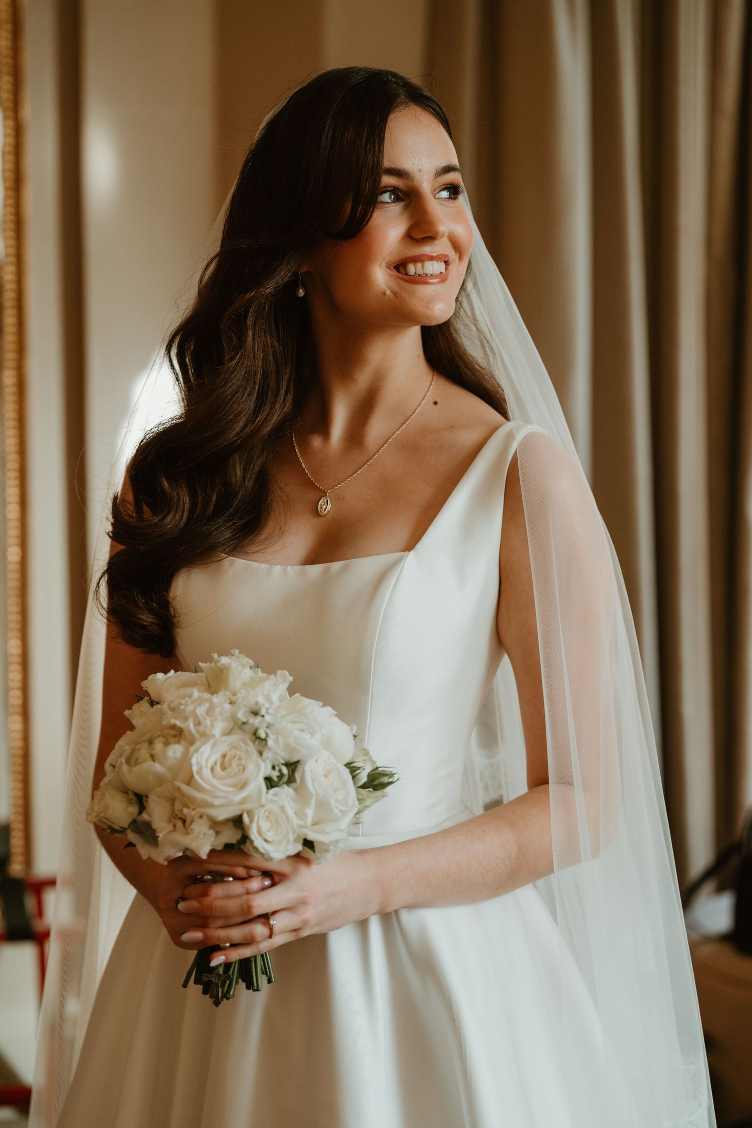 Beautiful bride in elegant white square neck wedding dress holds white rose bouquet while smiling warmly.