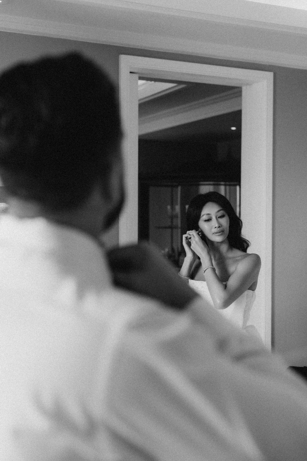 Black and white photo of bride putting on earrings reflected in mirror while groom stands nearby.