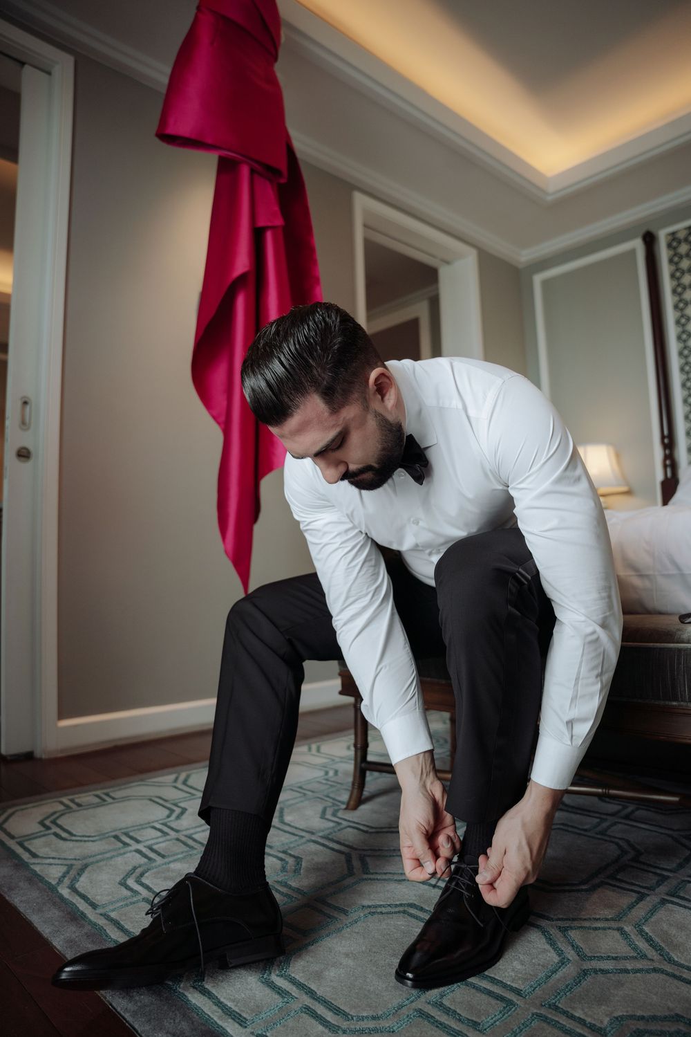 Groom in white shirt tying black dress shoes while sitting in elegant hotel room before wedding.