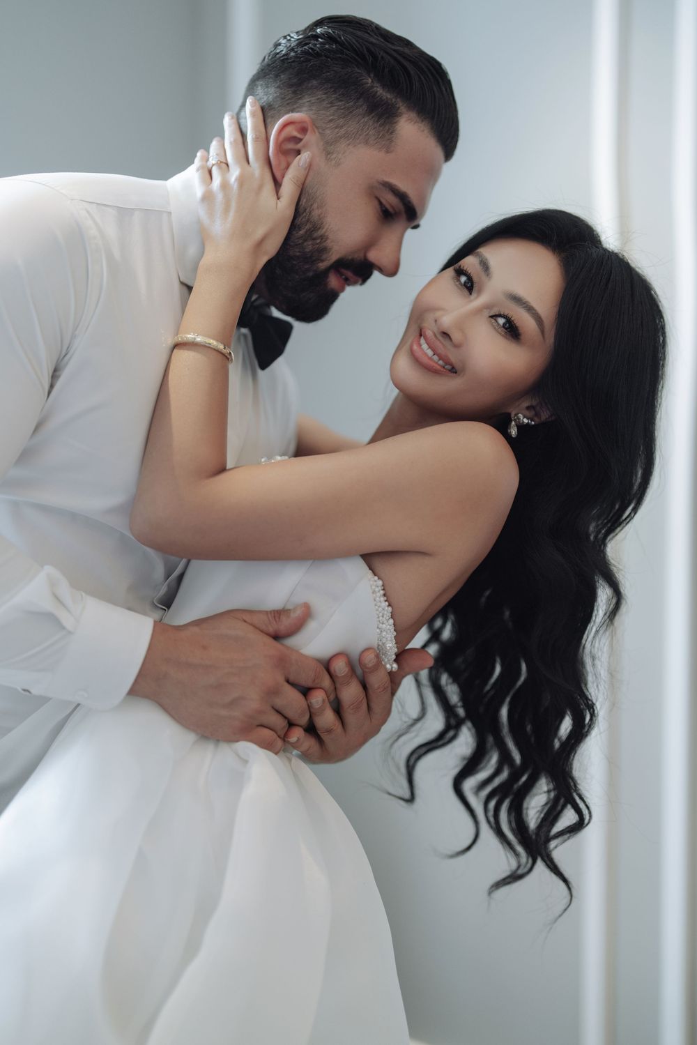 Interracial couple in wedding attire embracing tenderly, bride smiling in white gown.