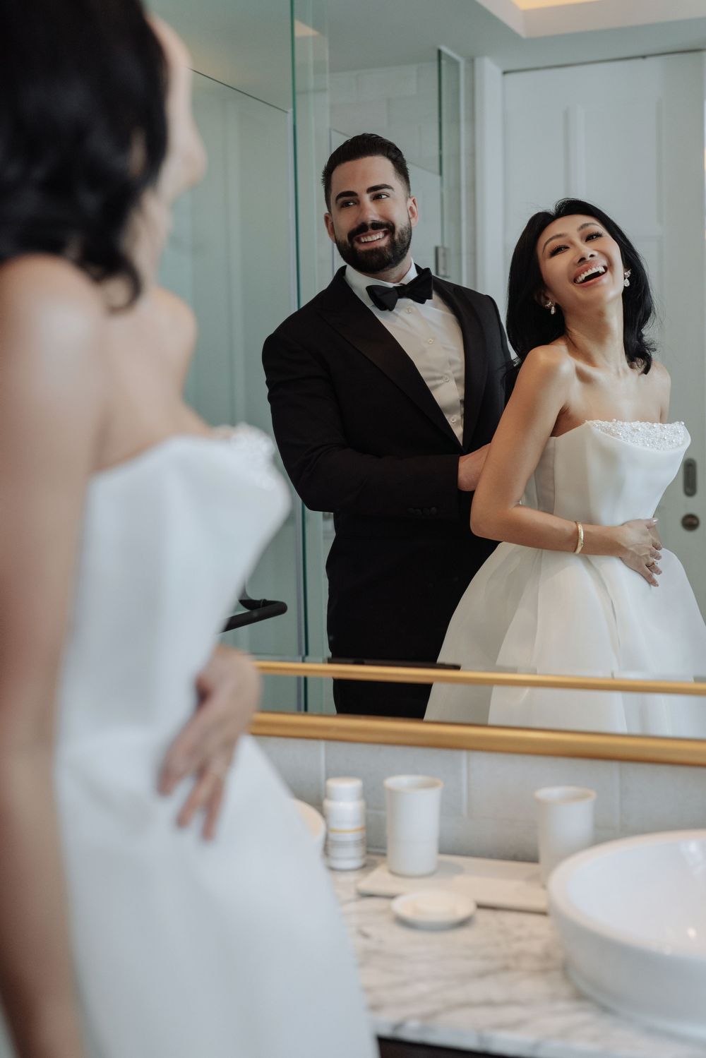 Smiling groom in tuxedo helping laughing bride with white dress in luxury bathroom mirror.