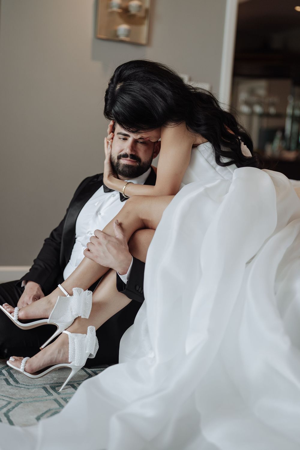 Groom in black tuxedo cradling bride in white gown on floor in intimate wedding moment.