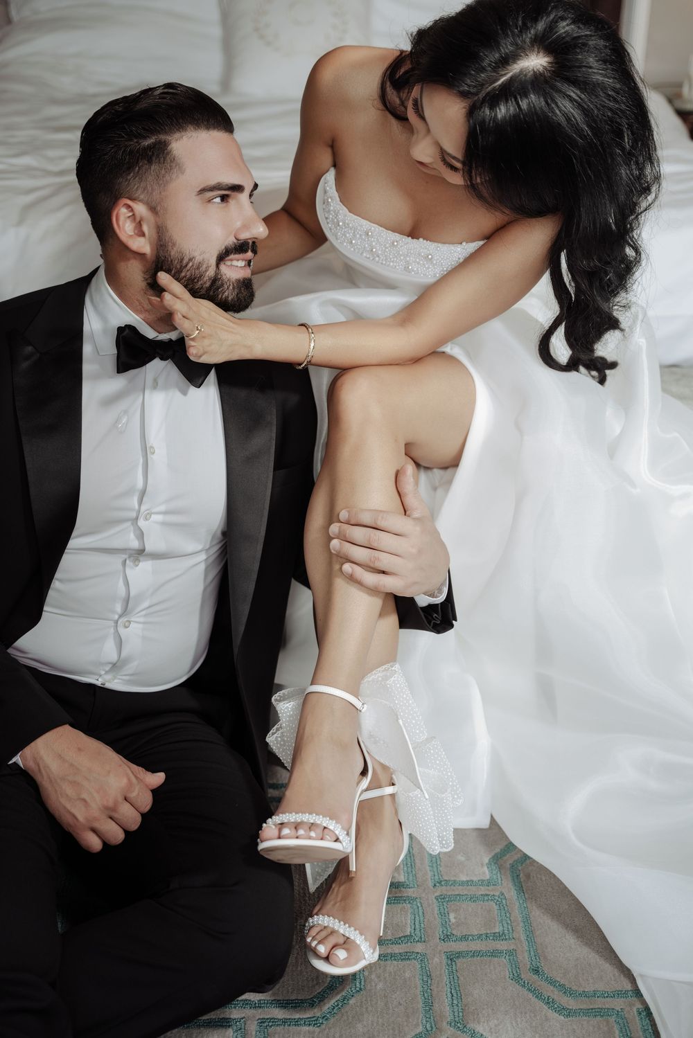 Groom in tuxedo sitting on floor as bride in white strapless gown tenderly touches his face.