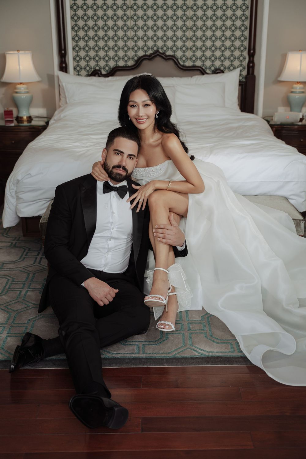 Bride and groom posing elegantly before ornate bed, she in white gown, he in black tuxedo.