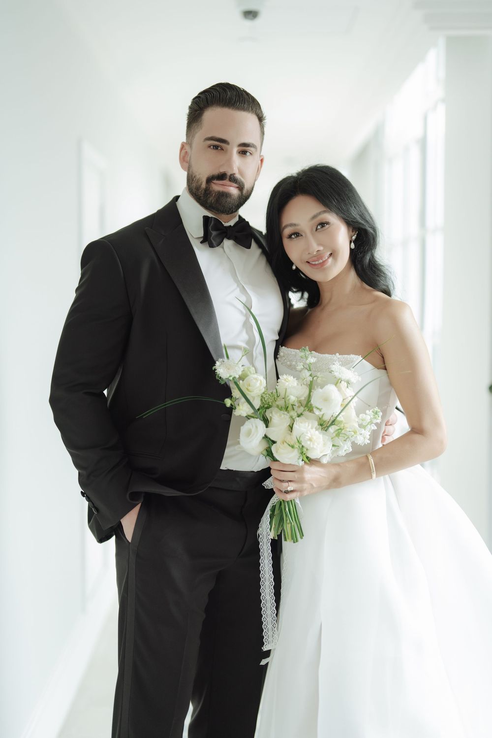 Bride holding white rose bouquet beside bearded groom in black tuxedo in bright hallway.