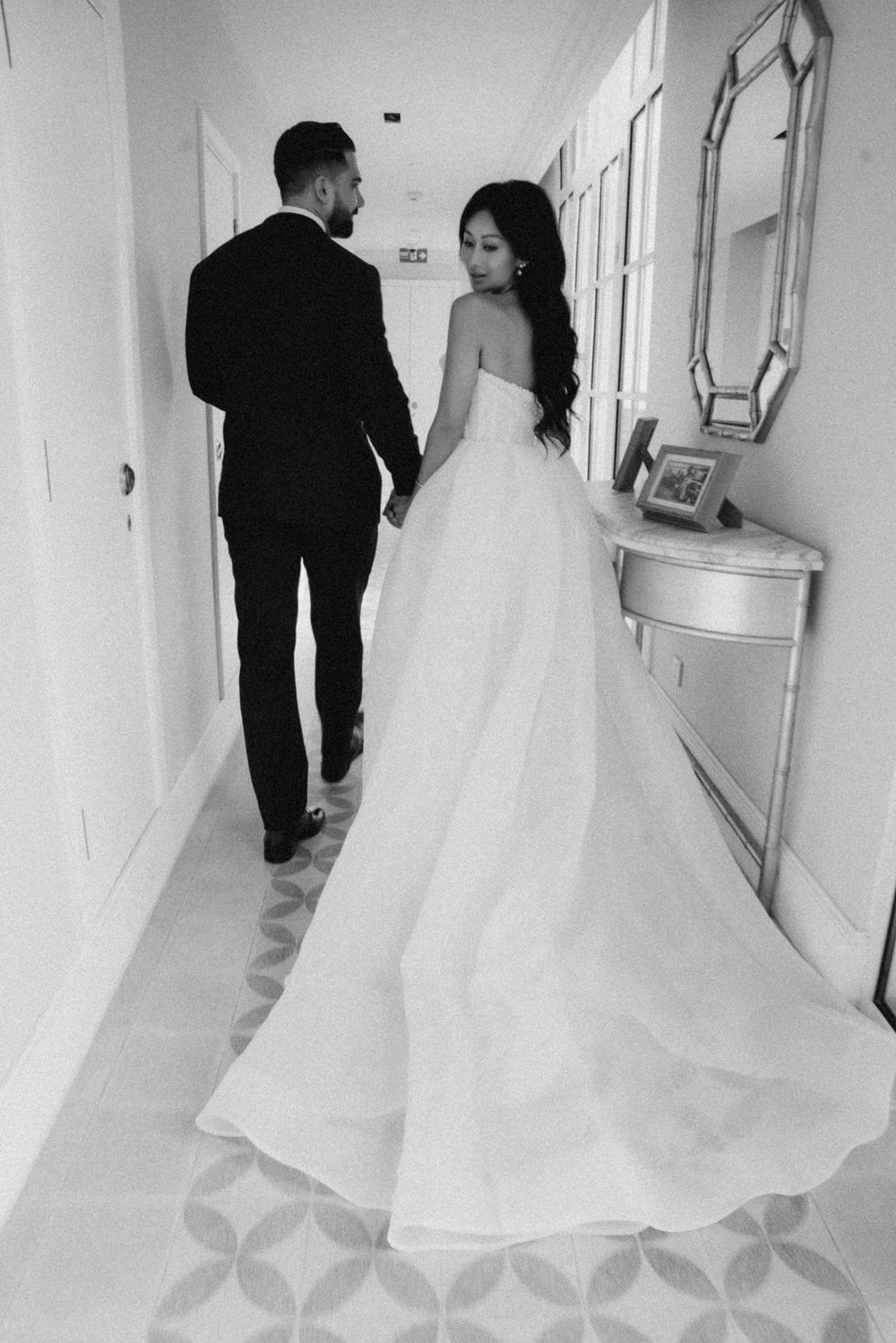Black and white photo of bride and groom walking hand in hand down elegant hotel corridor.