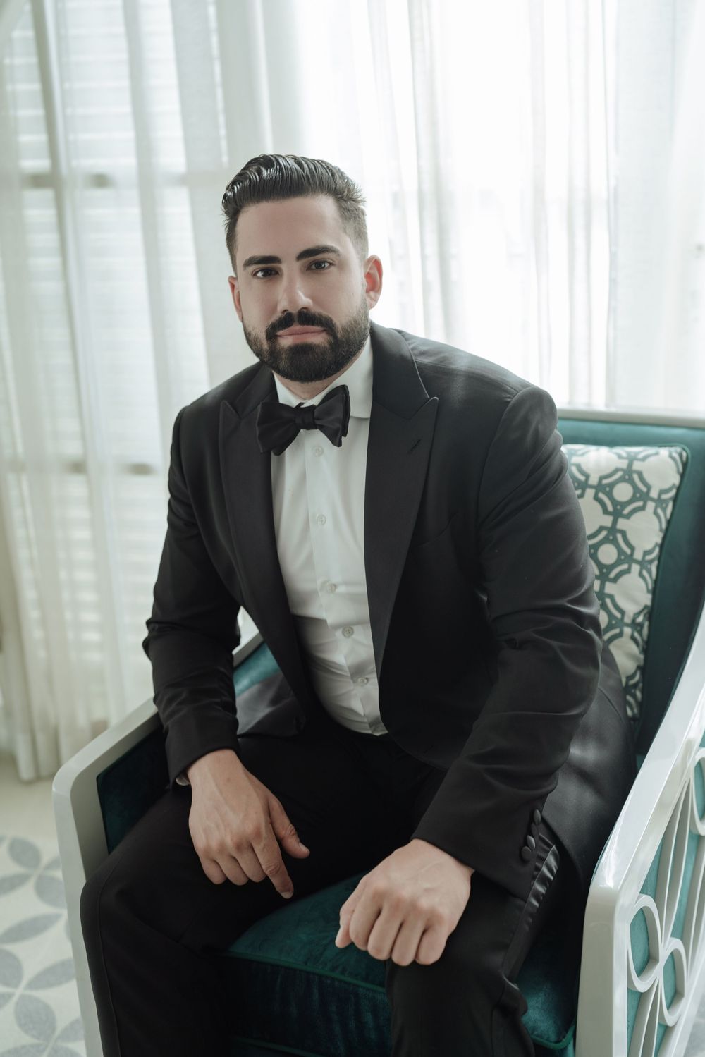 Bearded groom in black tuxedo with bow tie seated elegantly in a teal velvet chair by sheer curtains.
