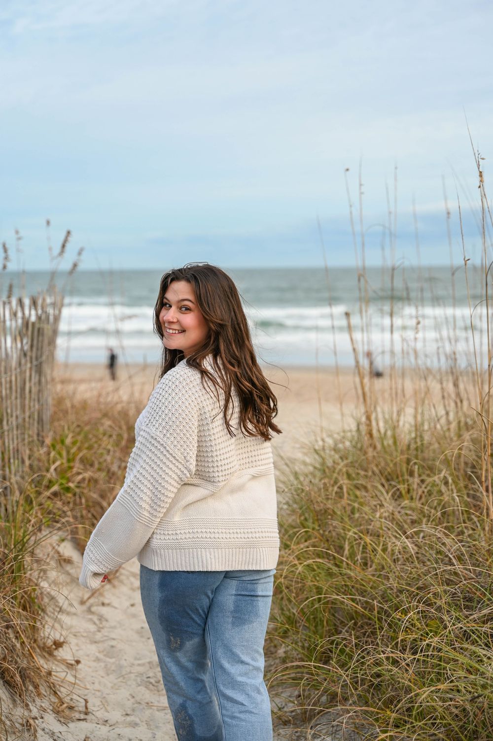 Woman in cream sweater and jeans stands on sandy beach path with sea and dunes in background on cloudy day.