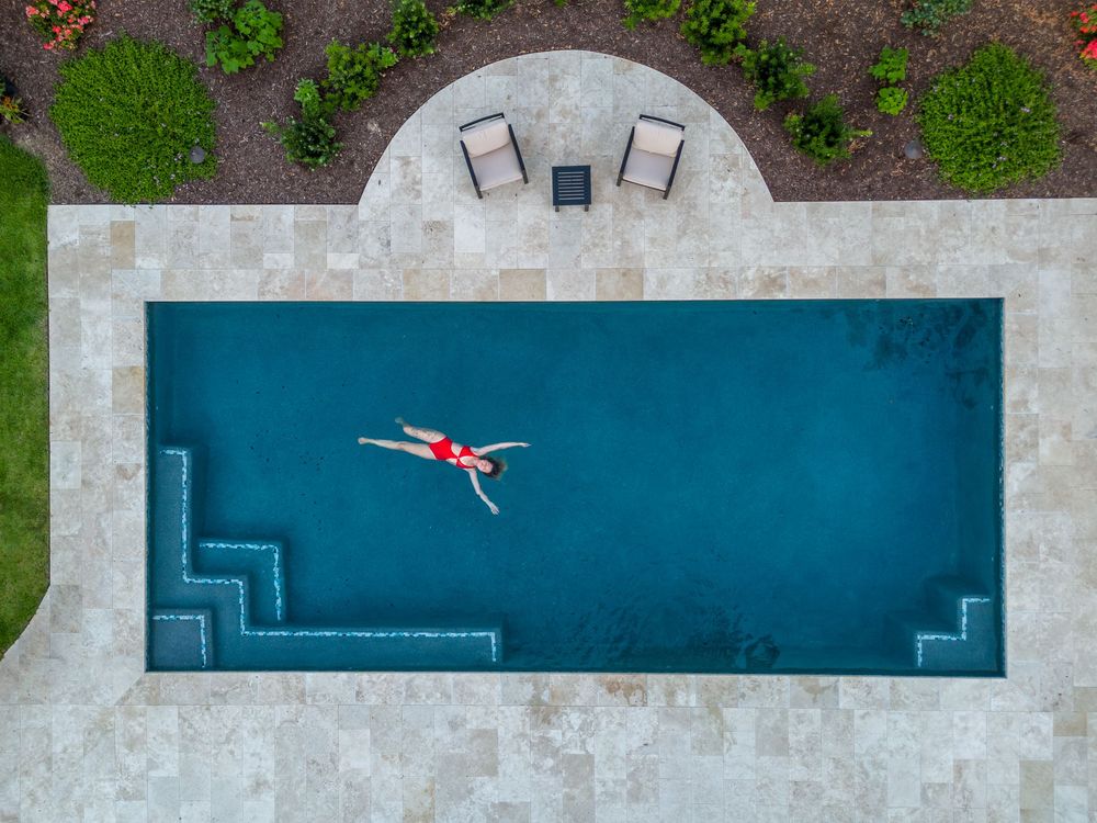 Aerial view of a rectangular swimming pool with turquoise water surrounded by concrete decking and lush greenery.