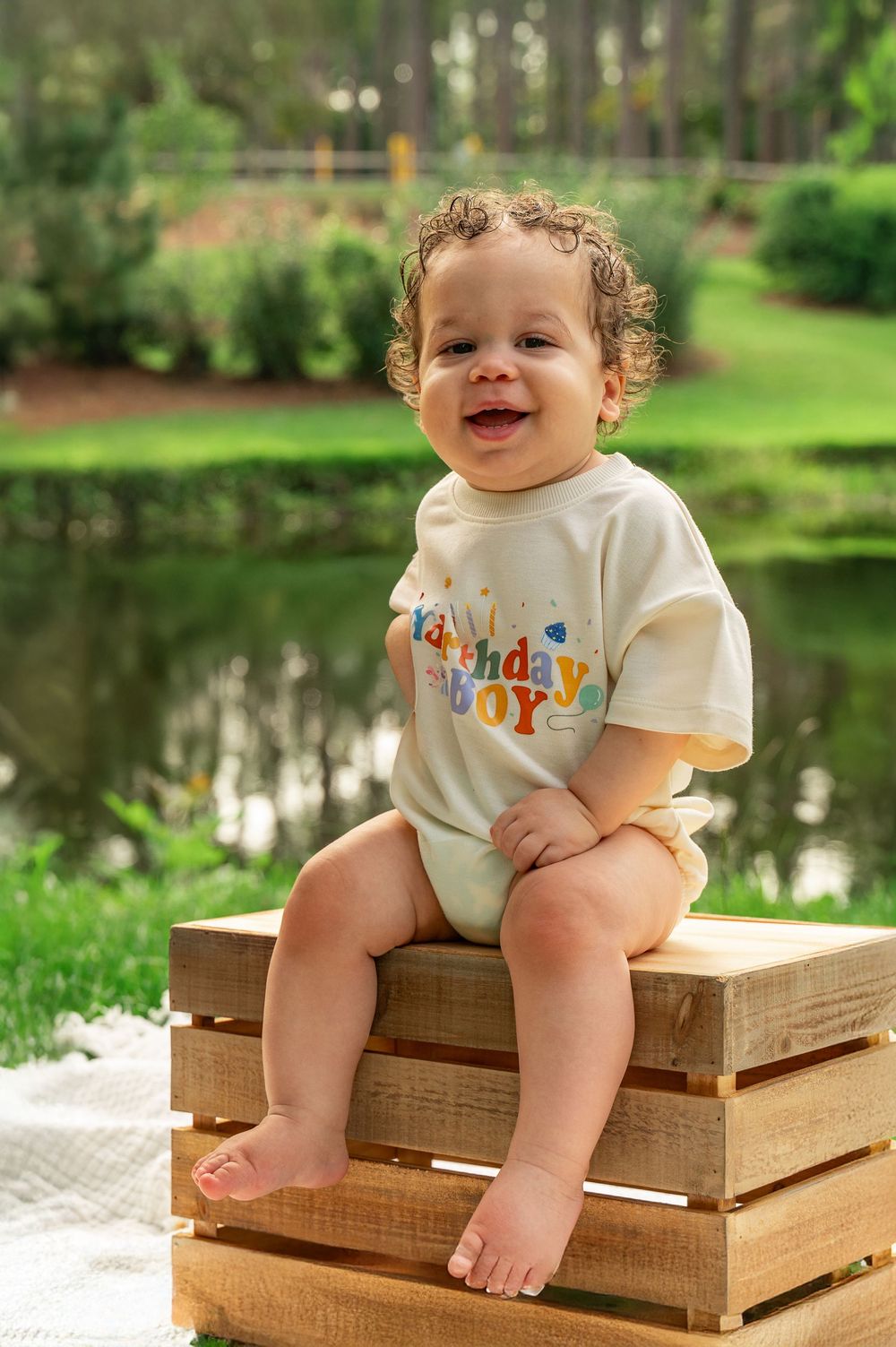 A small child in a white onesie sits on wooden crates outdoors near a pond on a sunny day.
