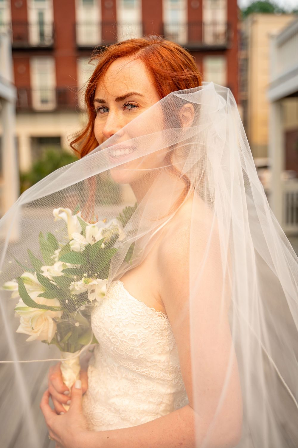 A bride in a white lace gown and flowing veil holds a bouquet of white flowers on her wedding day.