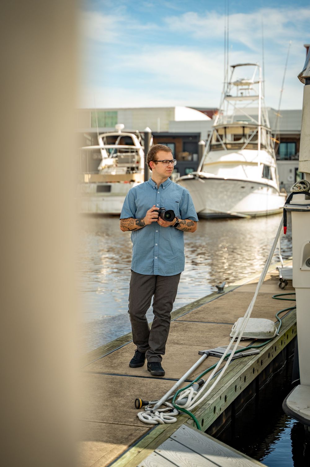 Photographer in blue button-down shirt taking photos on a wooden dock with boats in the marina background.