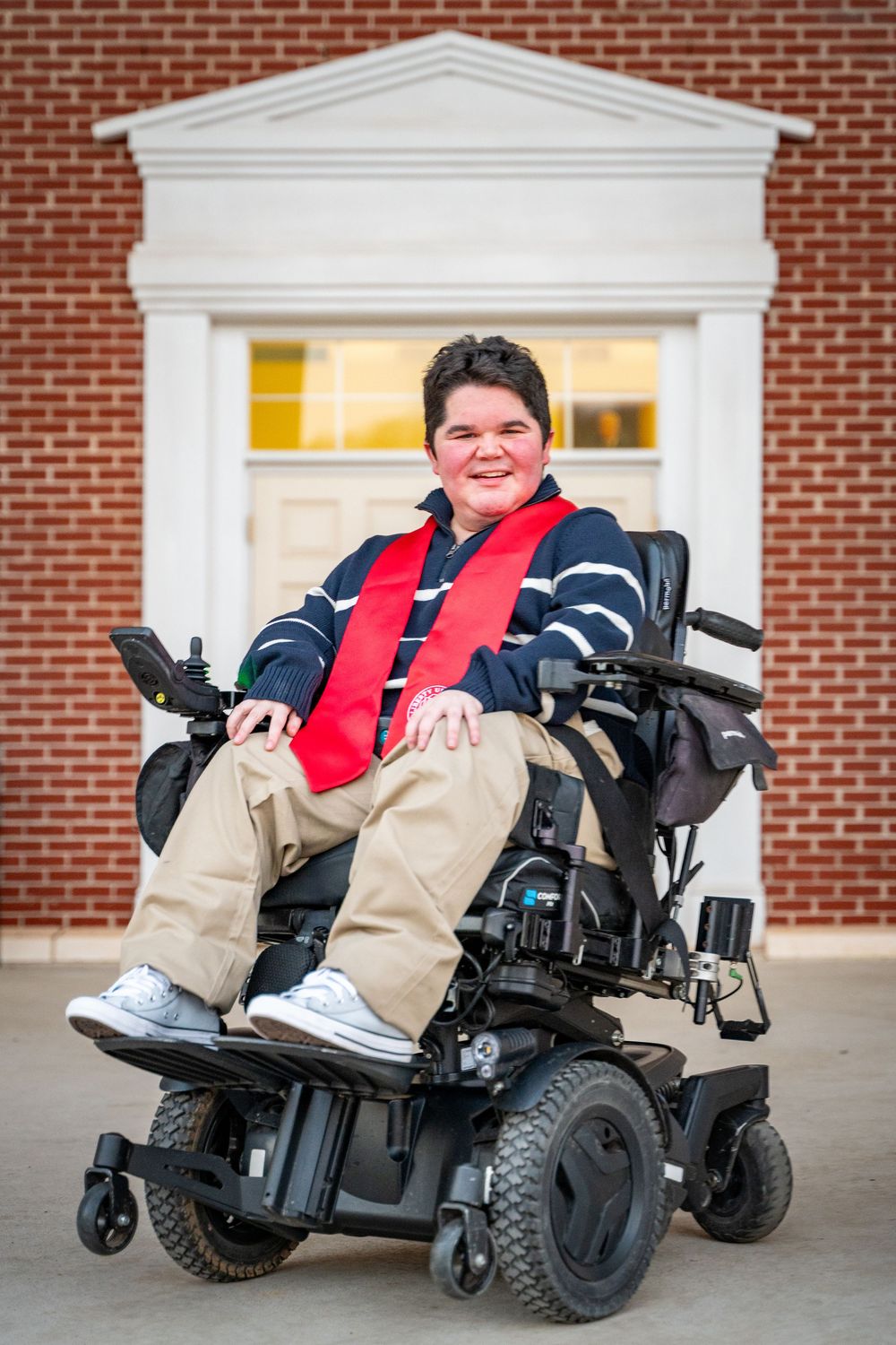 A person in a navy striped sweater and red scarf sits in a power wheelchair in front of a brick building entrance.