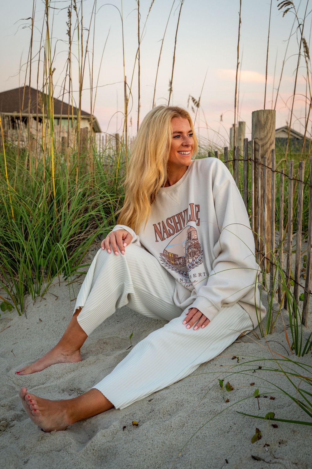 Casual beachwear photo featuring someone in a white Nashville sweatsuit relaxing in coastal sand dunes at dusk.