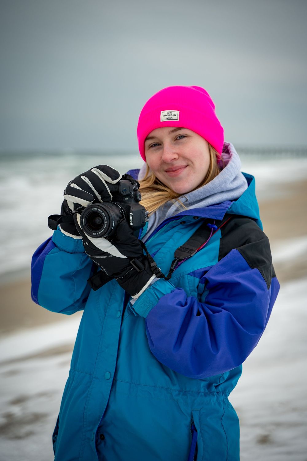 A photographer in a bright blue winter coat and hot pink beanie takes photos on a cloudy beach.