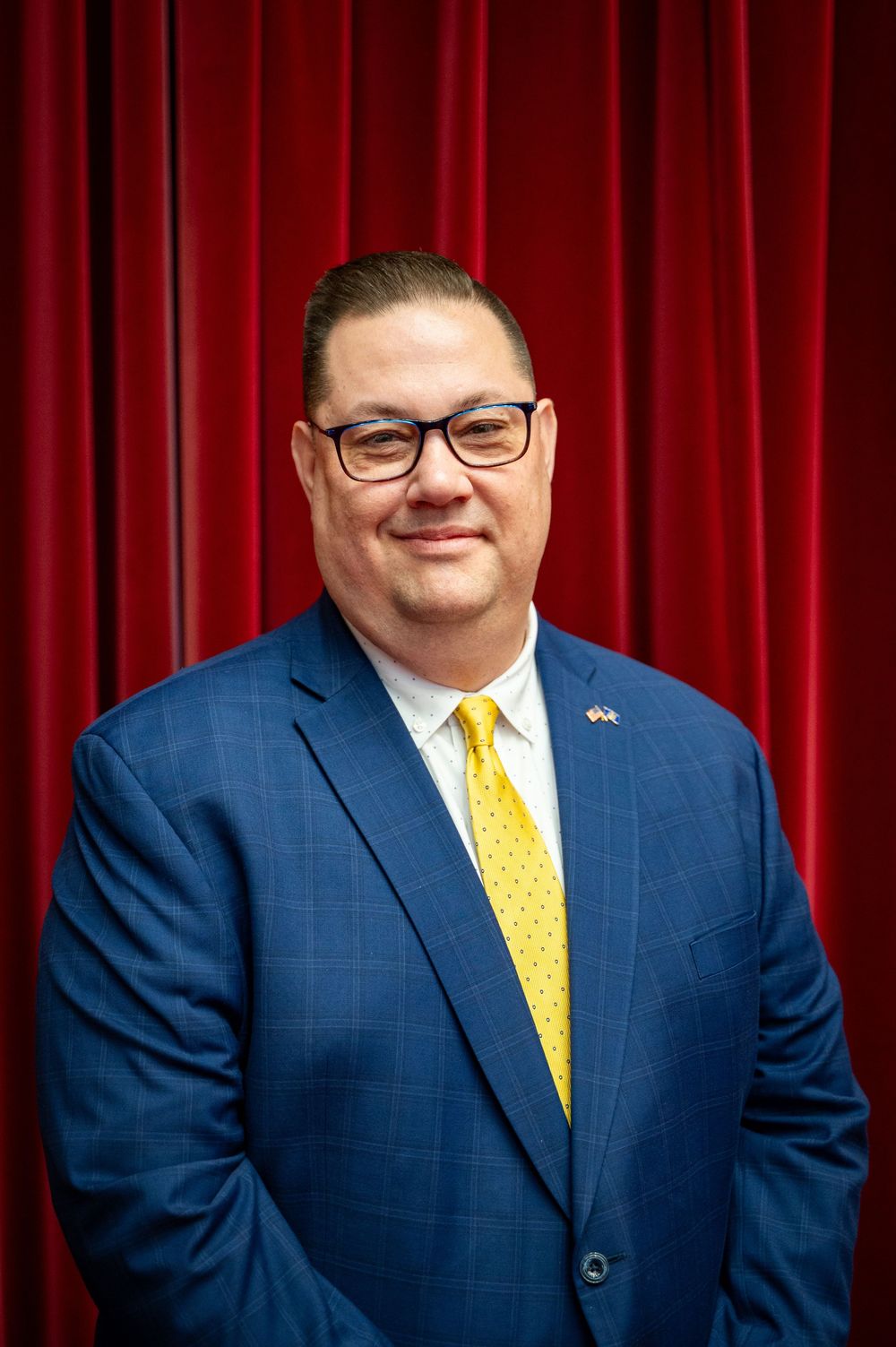 Professional headshot portrait of a person in a blue suit and yellow tie standing in front of a red curtain backdrop.
