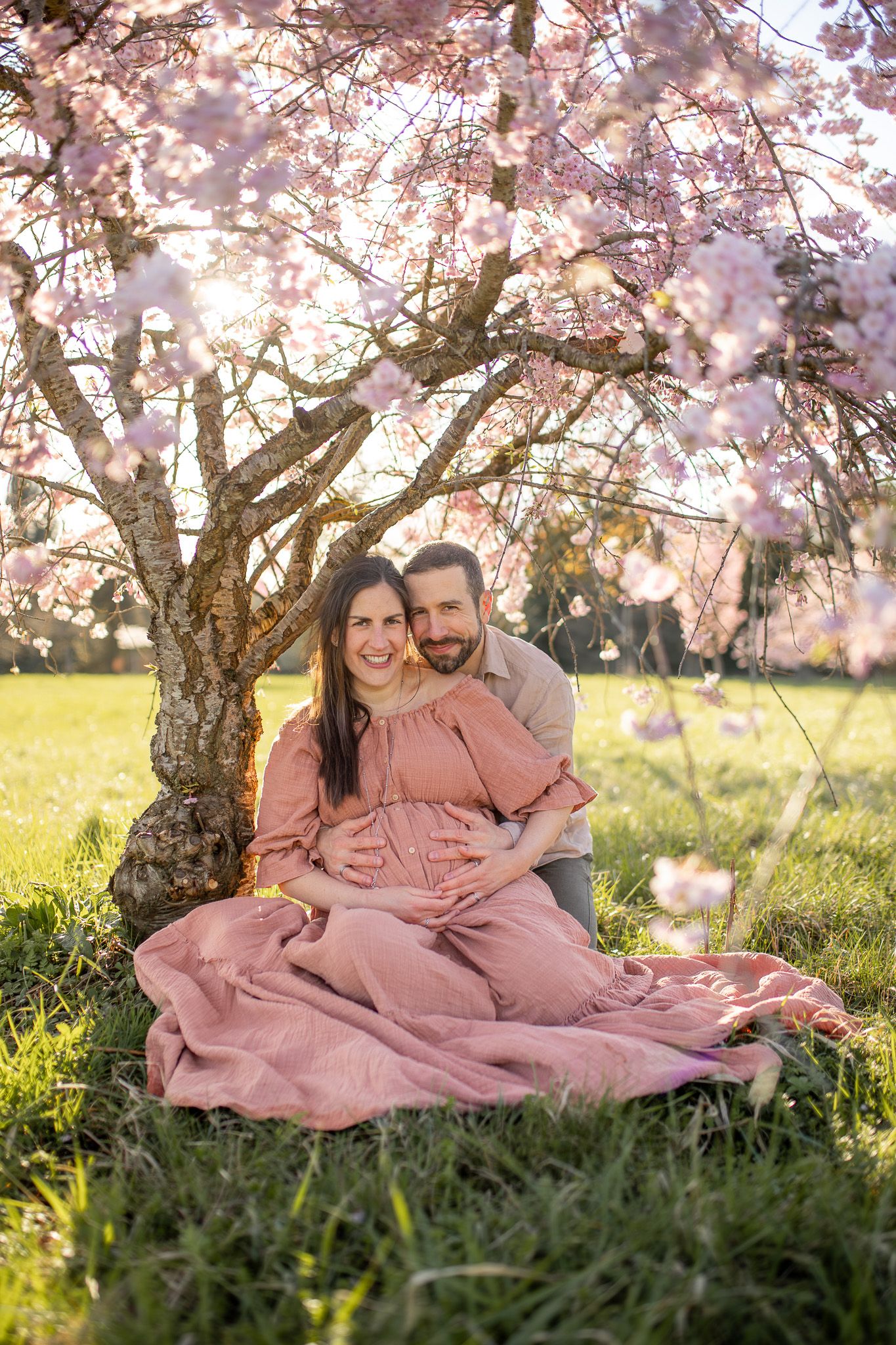 Couple souriant regard caméra lors d’une séance grossesse sous les cerisiers en fleurs