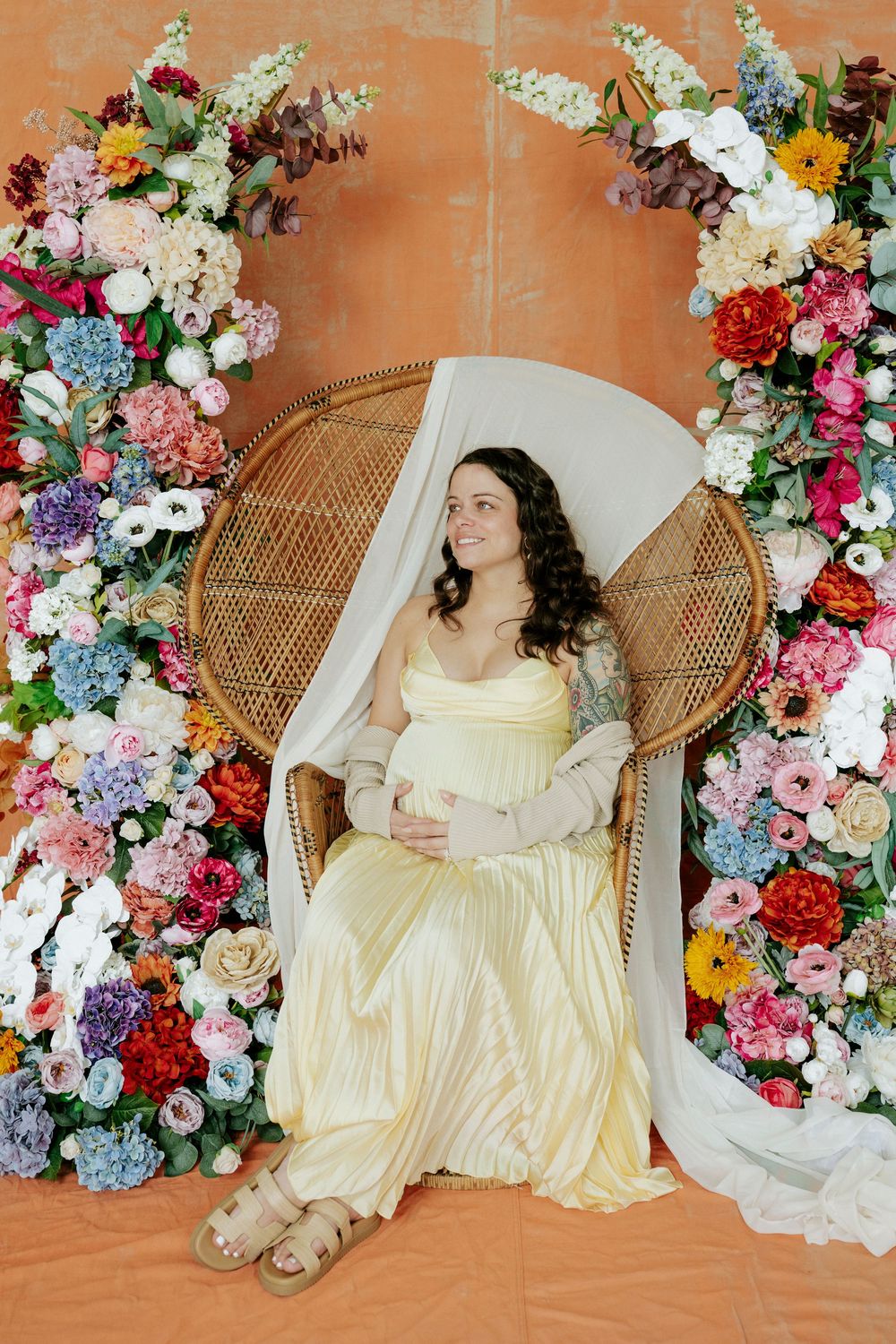 A floral maternity photoshoot featuring a wicker peacock chair surrounded by colorful blooms against a terracotta backdrop.