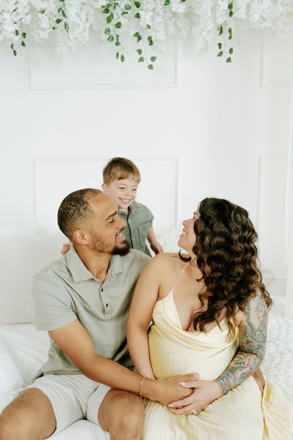 A couple shares intimate moments while sitting together on a white couch with greenery decor in the background.