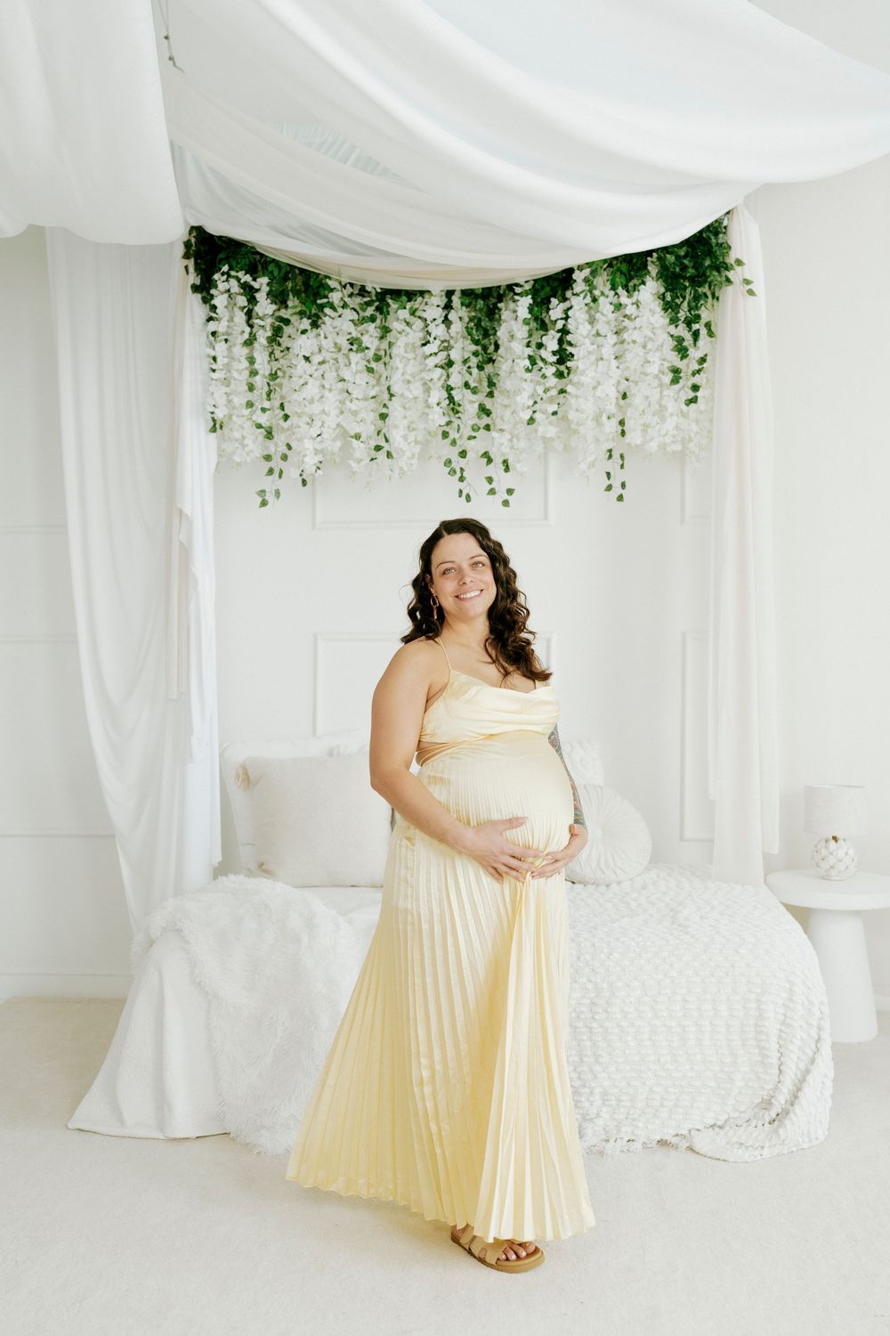 A maternity photo featuring a pleated cream dress against white drapes with hanging greenery decor.