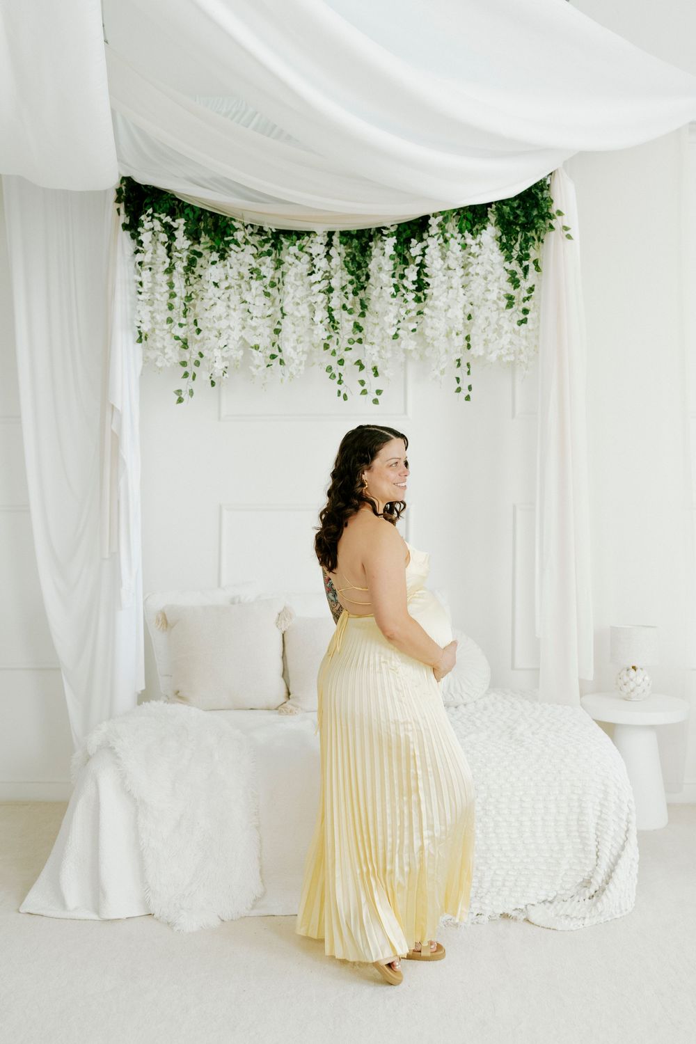 A romantic maternity photo series in a dreamy white studio setting with draped fabric and hanging greenery.