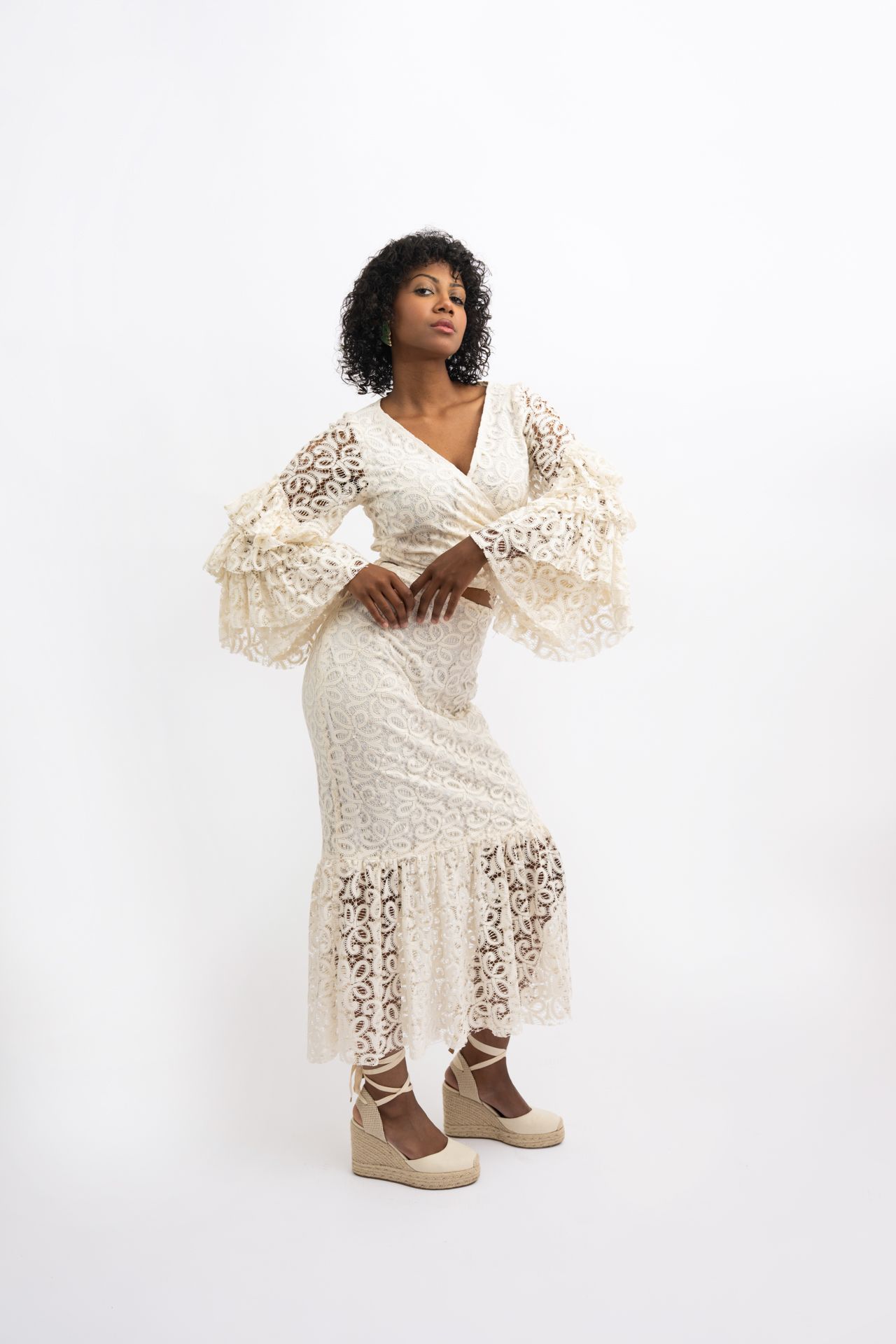 Woman in cream lace midi dress with bell sleeves and wedge espadrilles posing against white background.