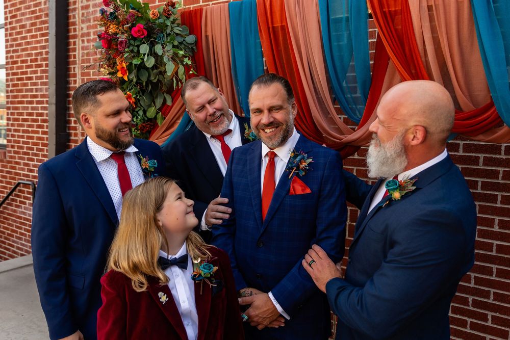 Groom and groomsmen in navy suits with red ties laugh together at colorful wedding ceremony backdrop at Founders One Nine in Omaha, NE.