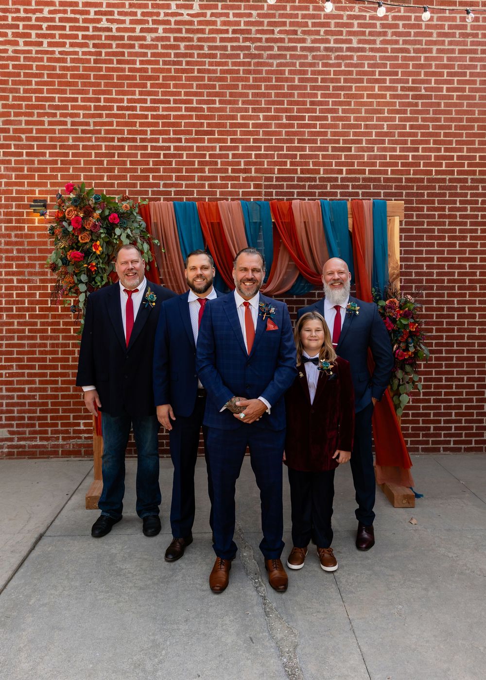 Five groomsmen in navy suits pose before a colorful draped backdrop with floral arrangements at a wedding at Founders One Nine in Omaha, NE.