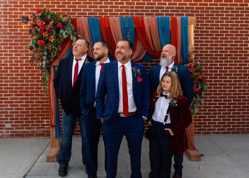 Wedding party in navy blue suits with red ties posing before a floral arch against a brick wall at Founders One Nine in Omaha, NE.