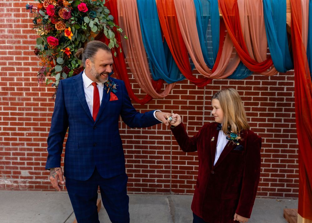 Groom in blue plaid suit and boy in burgundy jacket hold hands in front of colorful draped brick wall at Founders One Nine in Omaha, NE.