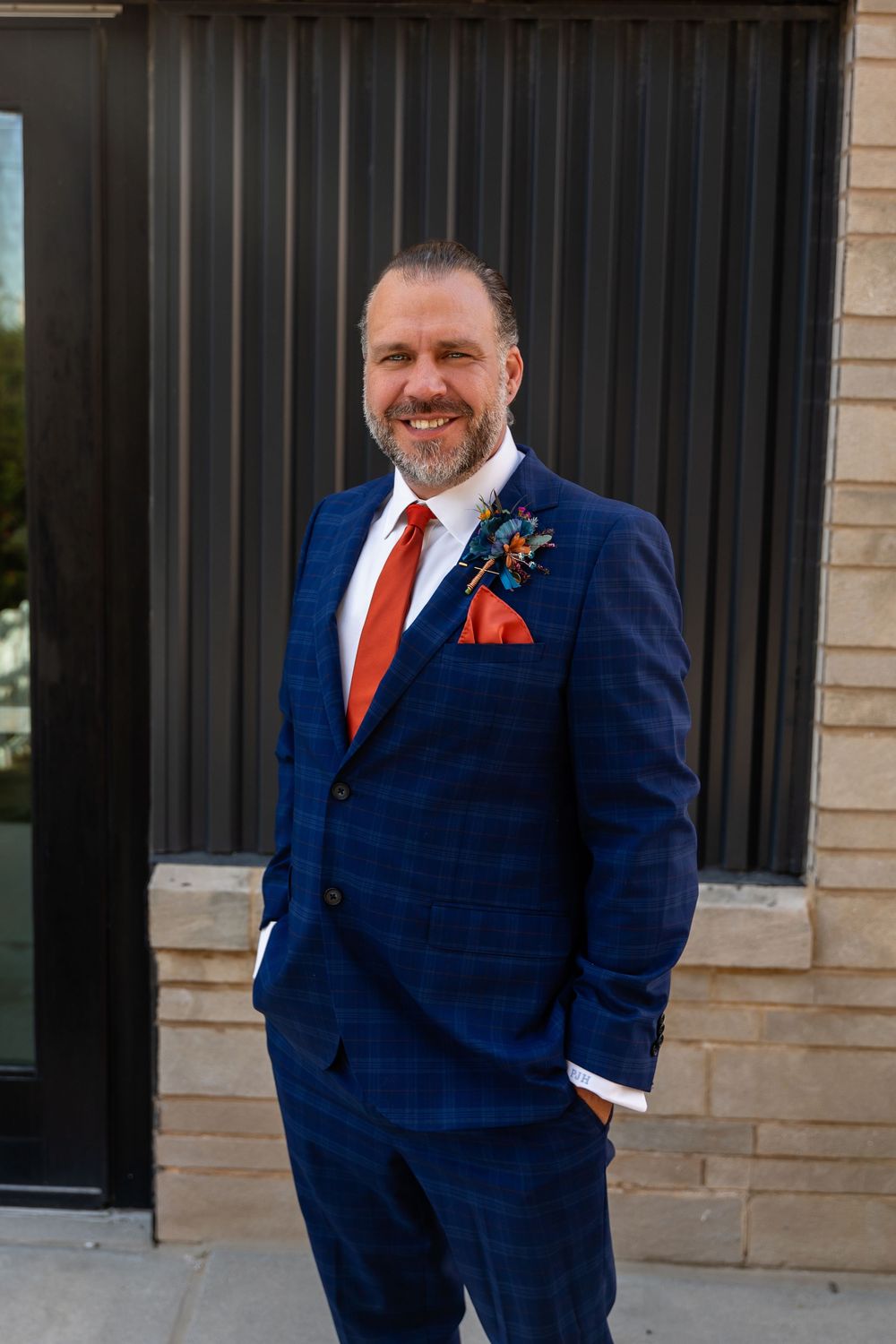 Groom in navy plaid suit with orange tie and boutonniere smiles confidently outdoors at his wedding at Founders One Nine in Omaha, NE.