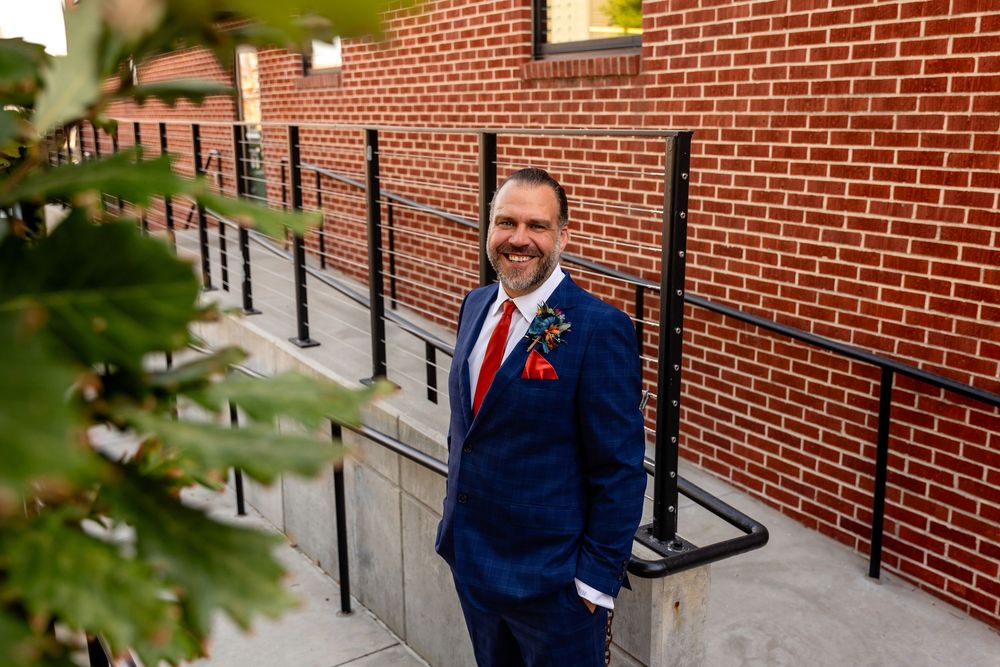 Smiling man in blue suit with red tie and boutonniere standing by brick wall outdoors at Founders One Nine in Omaha, NE.